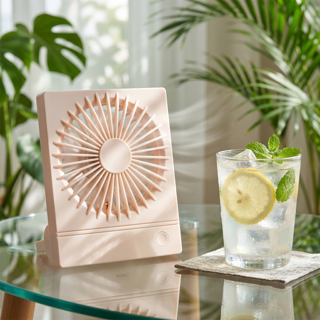 A pink ultra-thin personal desk fan on a glass table next to an iced lemon water glass with green plants in the background.