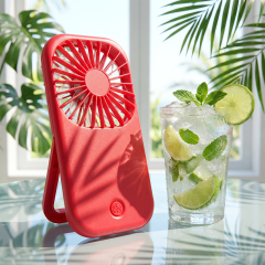 A bright red foldable pocket fan standing upright on a glass table next to a refreshing iced lime and mint drink.