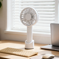 A white handheld mini fan standing in its vertical base on a wooden desk next to a laptop, keyboard, and mouse in front of a sunlit window with blinds.