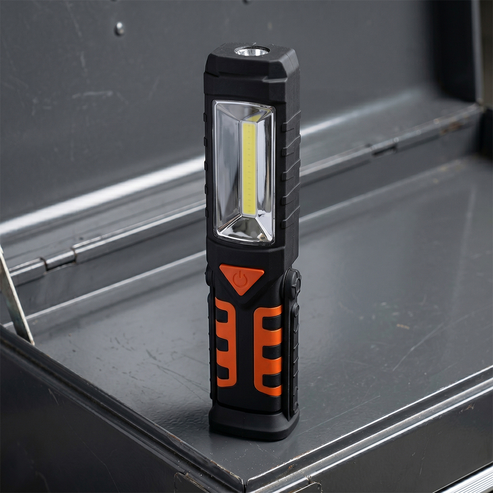 An orange and black magnetic LED work light standing vertically on a dark grey workbench inside a garage.