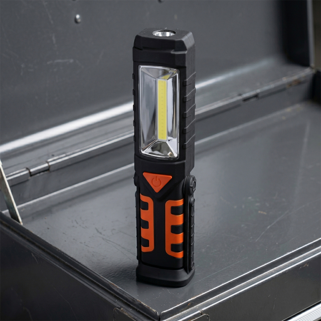 An orange and black magnetic LED work light standing vertically on a dark grey workbench inside a garage.