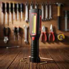 A black and red multifunctional LED work light standing on a wooden workbench with tools in the background.