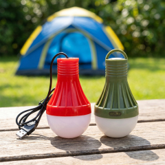 A red USB-powered bulb light and a green battery-powered bulb light standing side by side on a wooden outdoor bench with a blurry blue tent and green grass background.