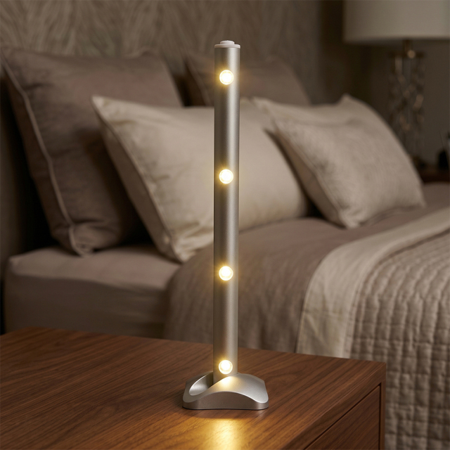 A silver LED light bar standing vertically on a wooden nightstand next to a bed, emitting warm light in a dimly lit bedroom.