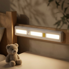 A wireless COB LED light bar mounted on a wooden bed headboard illuminating a nightstand next to a small plush teddy bear.