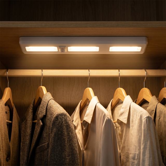 A wireless COB LED light bar installed under a shelf inside a dark wardrobe, brightly illuminating coats and shirts hanging below.
