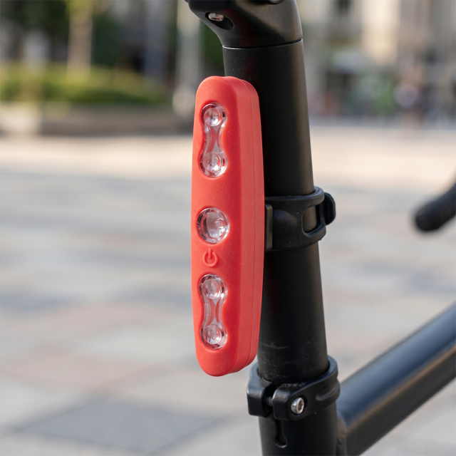 A red vertical LED bicycle tail light installed on a black metal seat post, providing rear visibility with a blurred outdoor background.