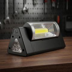 A black triangular magnetic COB LED work light resting on a wooden workbench, with a blurry workshop background showing tool pegboards.