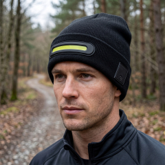 A close-up side view of a man outdoors wearing a black knitted beanie with a horizontal illuminated COB LED light strip on his forehead.