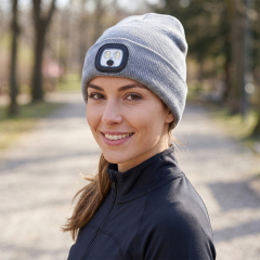 A smiling woman on a path wearing a classic grey winter beanie hat fitted with a compact square LED headlight.