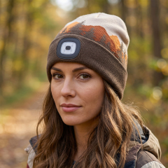 A woman in a forest trail wearing a brown knitted beanie with mountain patterns and an integrated square LED headlamp.