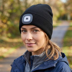 Portrait of a smiling woman outdoors wearing a classic black winter beanie integrated with a square rechargeable 4-LED headlight module.