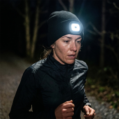 A woman jogging along a dark path at night wearing a black winter beanie with a bright illuminated LED headlamp.