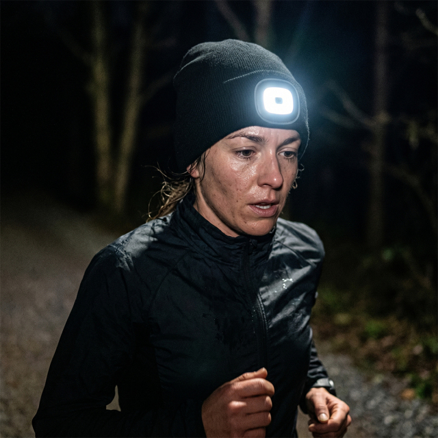 A woman jogging along a dark path at night wearing a black winter beanie with a bright illuminated LED headlamp.