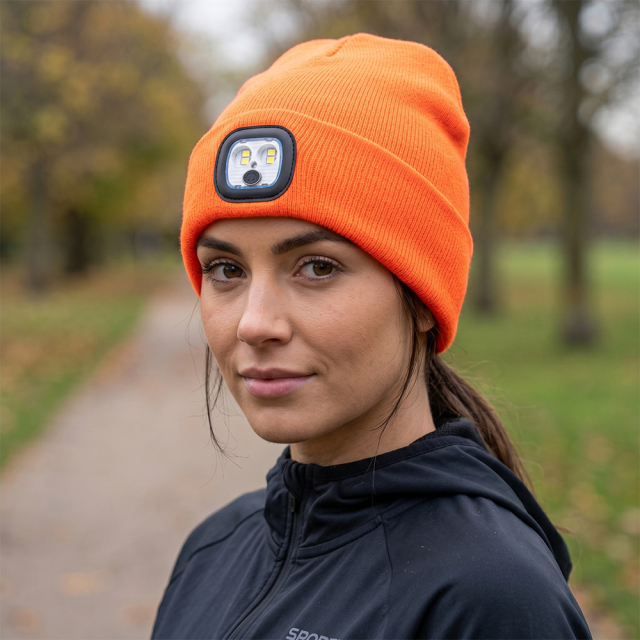 A woman wearing a high-visibility bright orange knitted beanie with an integrated square 4-LED headlamp on a park trail.
