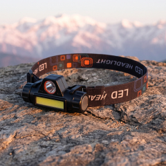 A compact plastic COB and XPE LED headlamp with a patterned elastic headband, sitting on a rugged rock face with a scenic mountain range background.