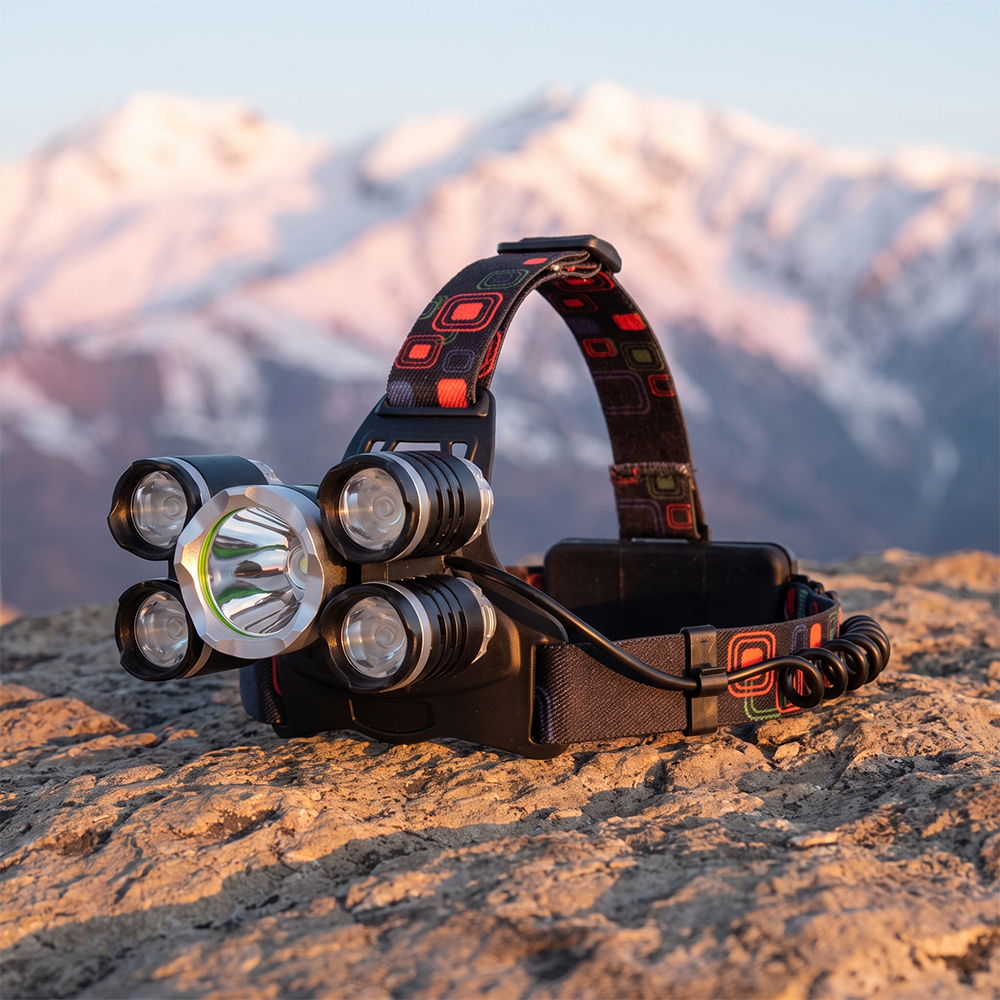 A real-world outdoor photo of a 5-LED high-bright headlamp sitting on a rugged, weathered rock face at dusk, with soft-focus snow-capped mountain peaks in the background.