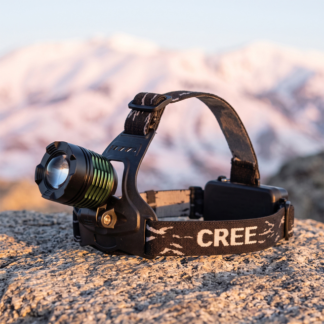 A 10W T6 LED headlamp with green cooling fins and a balanced three-strap headband, set on a rough rock with a soft-focus snowy mountain range background.