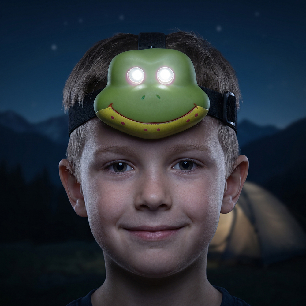 A real-world outdoor portrait of a smiling boy at night wearing a green frog-shaped kids LED headlamp that glows from its eyes, with a blurred dark camping background.