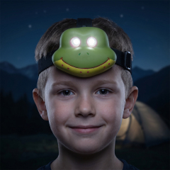 A real-world outdoor portrait of a smiling boy at night wearing a green frog-shaped kids LED headlamp that glows from its eyes, with a blurred dark camping background.