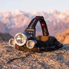 An outdoor photo of a multi-lens LED headlamp with a black adjustable three-strap headband, sitting on a flat rock with a soft-focus background of distant snowy mountains during sunset.