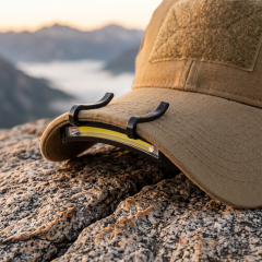 A real-world outdoor lifestyle photo of a black curved COB LED caplight clipped securely onto the brim of a tan tactical cap, resting on a rugged rock surface with hazy mountains in the background.