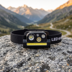 A real-world outdoor lifestyle photo of a black multi-LED work headlamp resting on a gray rocky surface, with a sweeping scenic mountain valley and winding path under soft sunlight in the background.