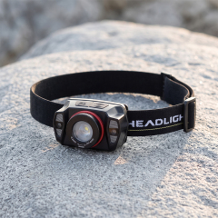 A real-world outdoor lifestyle photo of a black rechargeable headlamp with a distinctive red ring around its lens, placed on a light gray rock with natural textures under soft daylight.