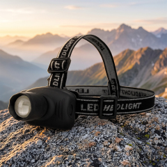 A real-world outdoor lifestyle photo of a compact black zoomable LED headlamp sitting on a rough stone surface, with a scenic background of misty valleys and distant mountain ranges at sunrise.