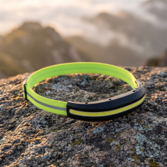 A real-world outdoor lifestyle photo of a black curved COB LED headlamp with a lime green high-visibility band resting on a textured gray rock surface, under warm sunrise light with mountains in the background.