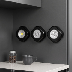 Three black rounded-square wireless LED cabinet lights mounted side-by-side on a dark gray wall above a white kitchen countertop with a mug and a coffee bean jar.