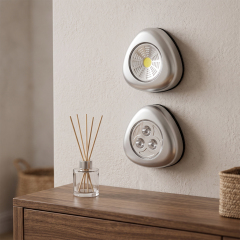 Two silver triangular wireless LED cabinet lights mounted vertically on a light-colored wall above a wooden side table with a glass aroma diffuser.