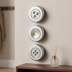 Three silver round wireless LED cabinet lights mounted vertically on a light-colored wall above a wooden table with an aroma diffuser.