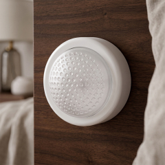 A white round wireless LED dome light mounted vertically on a dark brown wooden panel next to a bed.