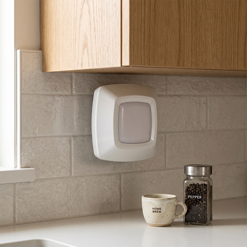 A real-world kitchen lifestyle photo showing a square white wireless LED puck light mounted vertically on a light gray tiled backsplash underneath wooden cabinets, next to a countertop with a coffee mug and spice jar.