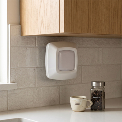 A real-world kitchen lifestyle photo showing a square white wireless LED puck light mounted vertically on a light gray tiled backsplash underneath wooden cabinets, next to a countertop with a coffee mug and spice jar.