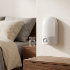 Studio view of an illuminated white arched LED night light mounted on a wall in a bedroom setting, located near a bed and a wooden nightstand.