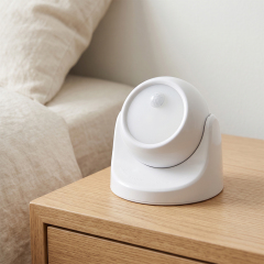 A white rotatable spherical wireless LED motion sensor night light sitting on a wooden bedside table next to a bed.