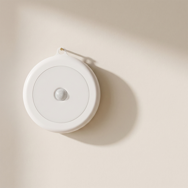 An overhead, three-quarter view lifestyle photograph showing the round white magnetic sensor light mounted above a simple shelf in a domestic hallway setting.