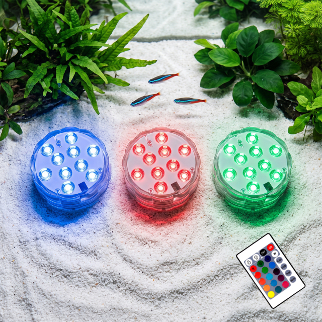 A scenic view of multicolored round submersible LED lights glowing in blue, red, and green on white sand inside an aquarium, with an IR remote control placed nearby.