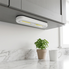 Studio view of an illuminated white slim LED light bar mounted under a grey kitchen cabinet, casting bright light down onto a marble countertop with a potted herb and a mug.