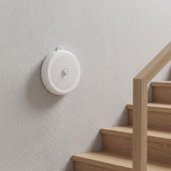 A scenic lifestyle photograph showing the round white magnetic sensor light mounted on a grey textured wall next to a wooden residential staircase.