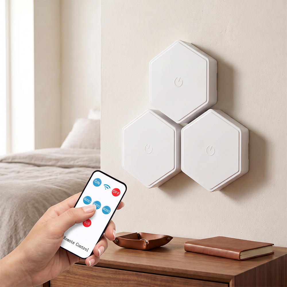 A person's hand holds a remote control pointing at a cluster of three unlit white hexagonal modular LED wall lights mounted on a bedroom wall. The scene is set above a wooden nightstand with a brown catchall tray and a book, with a bed visible in the soft-focus background.