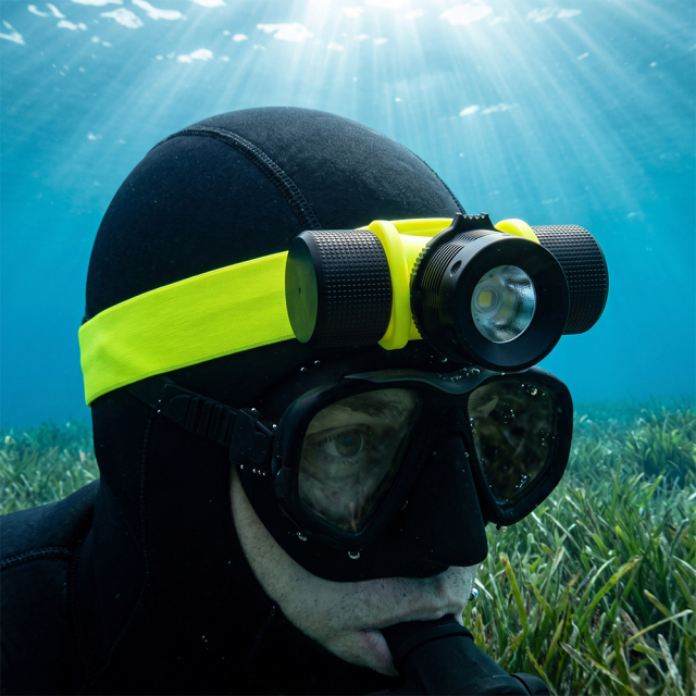 A close-up of a scuba diver wearing a black hood, mask, and a bright yellow professional waterproof LED diving headlamp underwater.