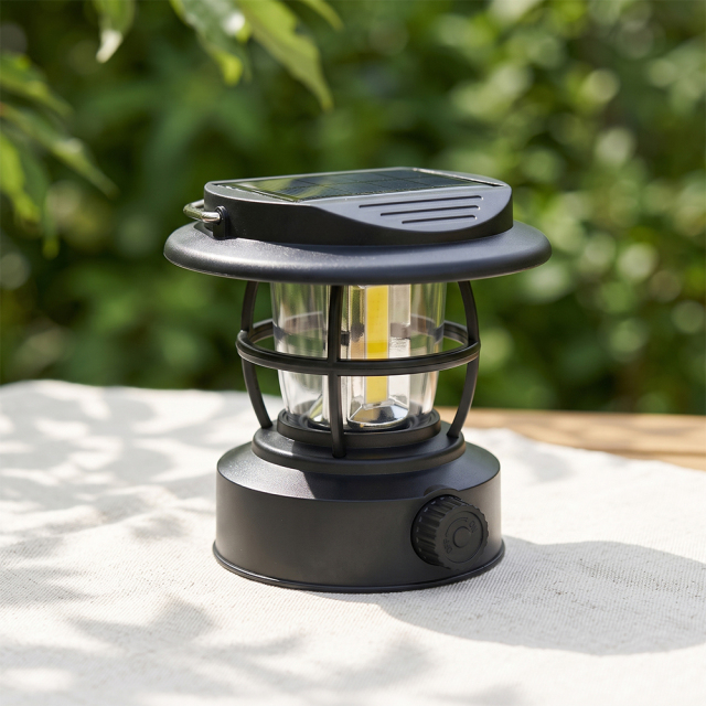 A black solar vintage camping lantern sitting on a light-colored outdoor table under bright daylight with lush green trees blurred in the background.