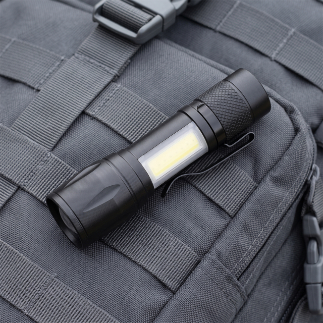 A compact black aluminum zoom flashlight with a metal pocket clip attached to a grey tactical backpack.