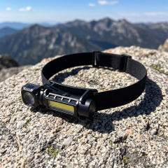 A black 3-in-1 LED headlamp mounted on an elastic strap resting on a mountain rock with a scenic outdoor background.