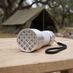 A silver 21 LED aluminum flashlight with a wrist strap lying on a wooden table in front of a camping tent outdoors.