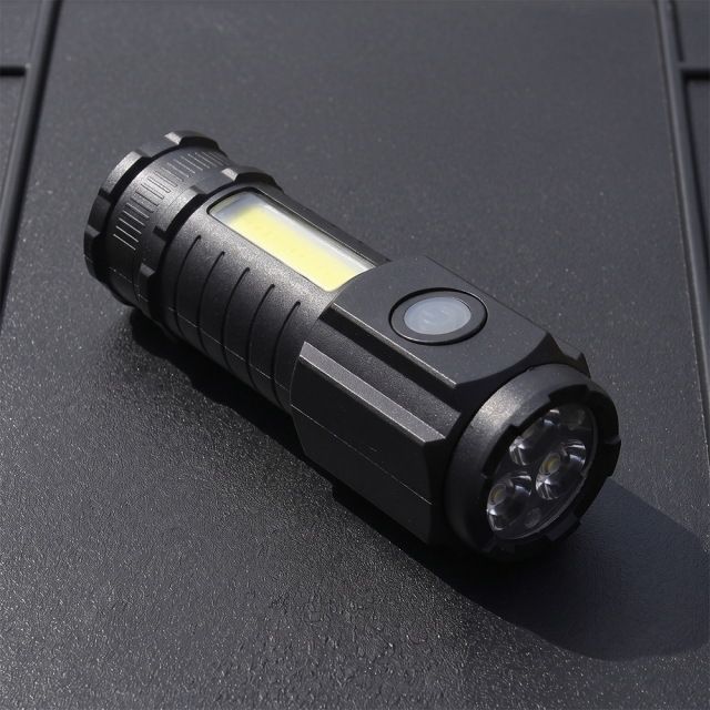 A black mini rechargeable flashlight with 3 LED front bulbs and a side COB floodlight panel lying on a dark surface.