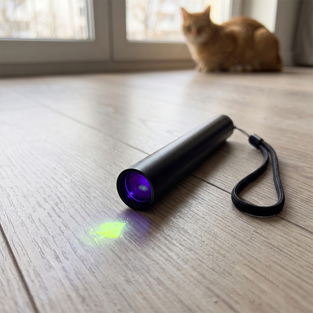 A portable UV blacklight flashlight illuminating a fluorescent stain on wooden flooring with a cat in the background.
